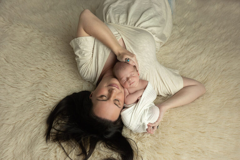 Mother lying on a cream rug, cuddling her sleeping newborn against her cheek.