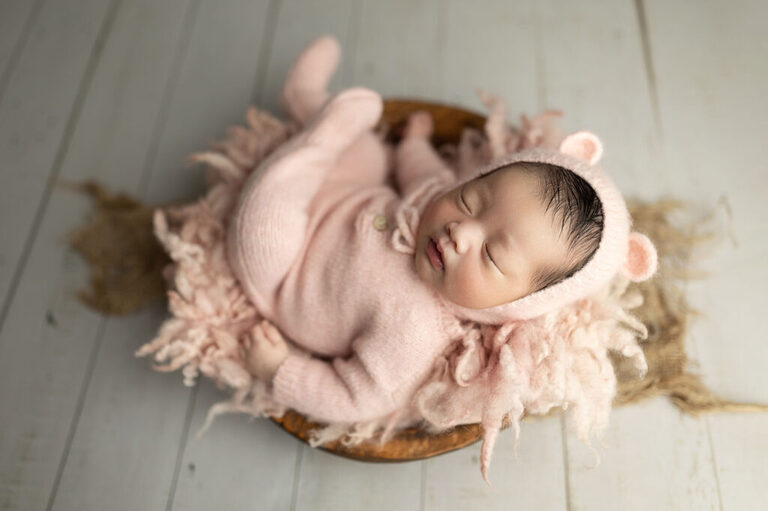 Sleeping newborn in a pink knitted outfit and bear bonnet, curled in a wooden bowl lined with soft pink fabric.