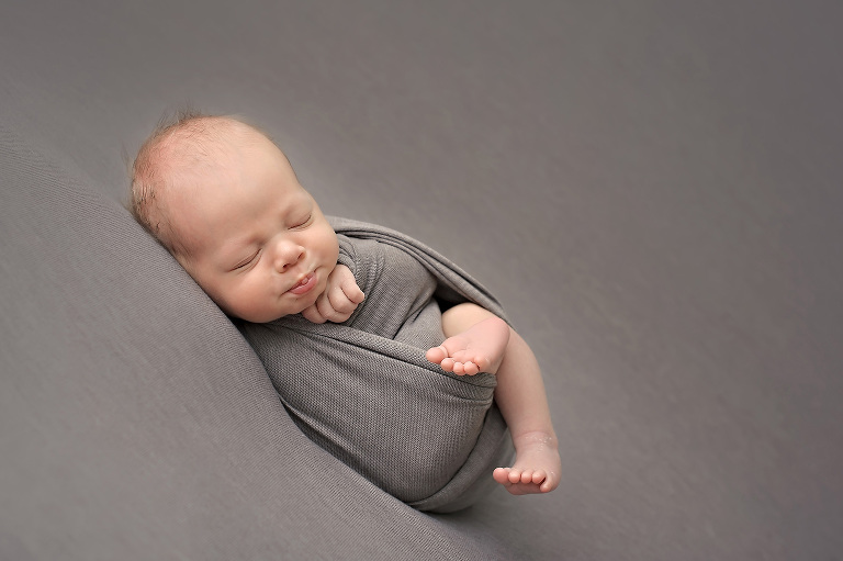 Newborn baby boy with pursed lips on gray stretch backdrop with matching gray stretch wrap