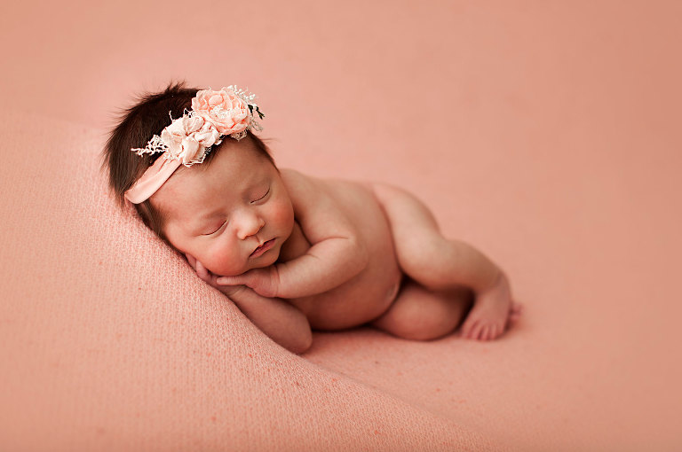 Newborn girl on peach background in side lying pose with peach headband by Golden Stitch Props