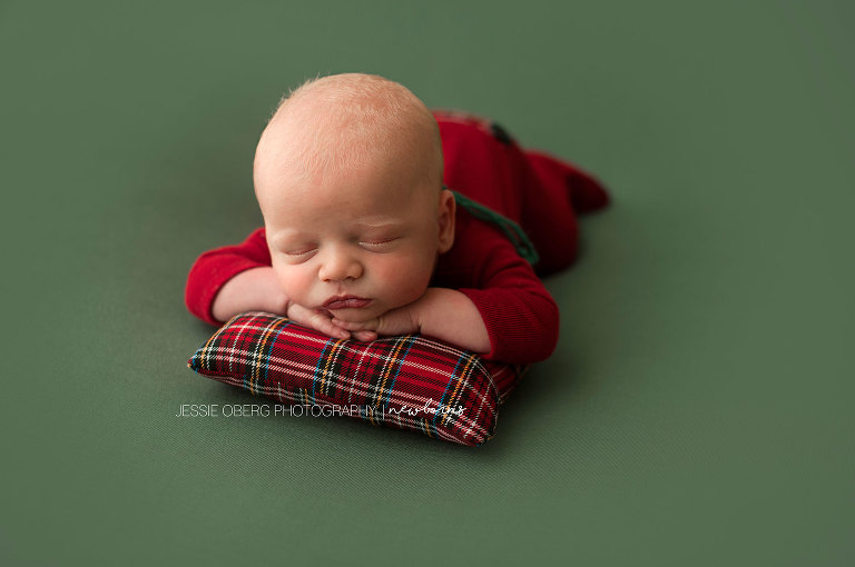 Newborn baby boy posed chin on hands on red plaid pillow and green backdrop. Such a cute red romper.