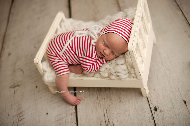 December newborn posed on tiny bed in red and white striped romper and bonnet.