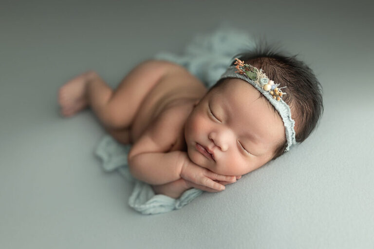 Sleeping newborn wearing a floral headband, resting on pale blue fabric with hands tucked beneath their cheek.