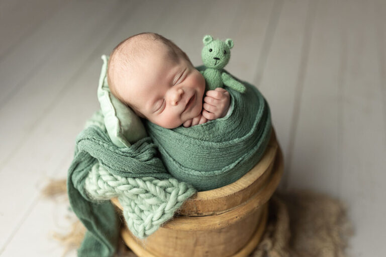 Smiling newborn wrapped in sage green fabric, holding a knitted bear while nestled in a wooden bucket.
