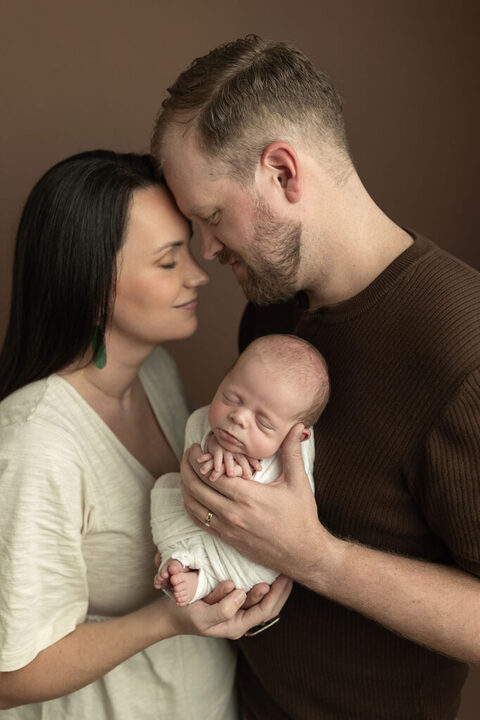 Parents touching foreheads while holding their sleeping newborn against a warm brown backdrop.