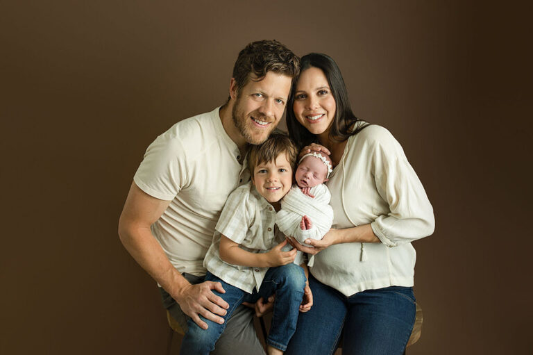 Parents and big brother cuddling their newborn together against a warm brown studio backdrop.